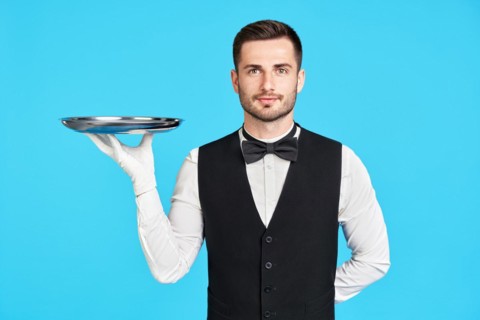 Attractive young waiter holding empty silver tray over blue background with copy space. Restaurant,