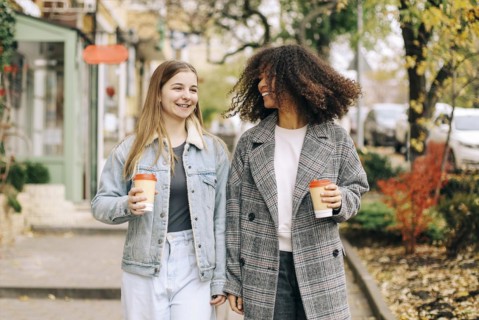 Two happy carefree multiracial girlfriends walking city street with take away coffee, talking and