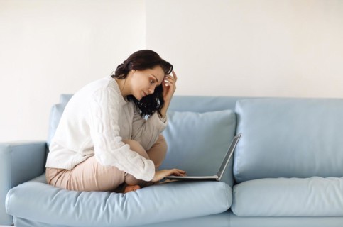 Young pensive woman in casual wear sitting with her legs on sofa using keyboard on laptop, staring