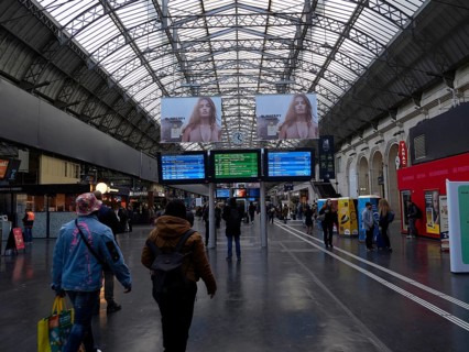 Travellers in a station concourse under a large glass-covered ceiling with timetable displays and