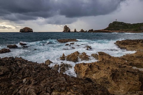 A rocky coastline with a pointed rock formation stretches into the sea at a bay, strong waves at a