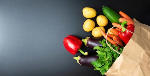 above view of fresh organic vegetables spilling out of paper shopping bag on dark kitchen counter,