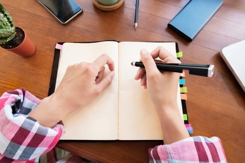 Young Caucasian attractive woman taking notes in her notebook while sitting in the kitchen at the