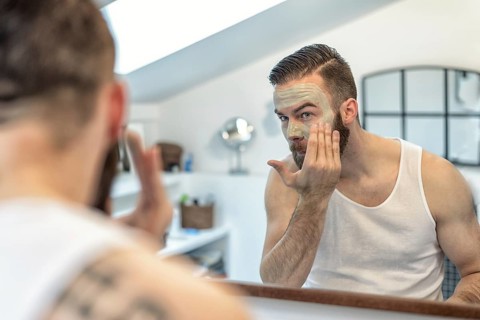 Reflection in the bathroom mirror of a young bearded man applying a face mask to his skin in a