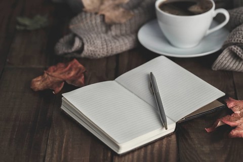 Notebook with pen, hot coffee cappucino, blanket and autumn leaves on old wooden background.