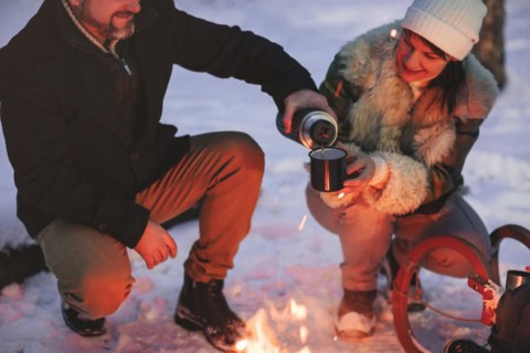 Joyful happy family couple in love basking by campfire outside in winter snowy forest drinking hot