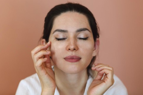 Positive young brunette female with eye patches touching her face and smiling during skin care