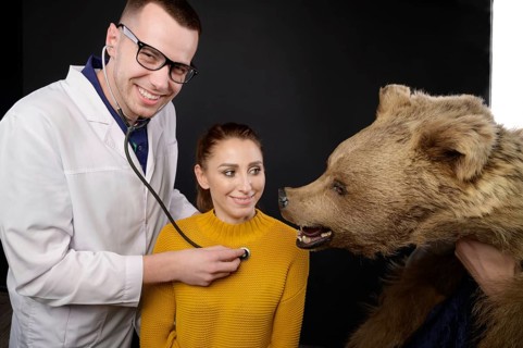 Smiling young doctor with stethoscope and patient examining bear, studio medical humor of whom to