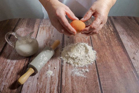 Woman breaking an egg, pouring it over flour to make a sponge cake on a wooden table with a rolling