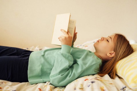 Caucasian blond teenage girl is lying on the bed and reading book. Reading fiction, hobbies,