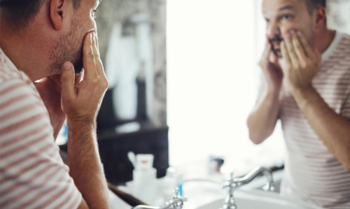 Millennial handsome man looking in mirror, applying moisturizing washing cream on cheeks in