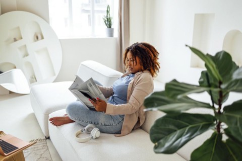 Photo of pretty happy young african woman sitting on sofa indoors at home while reading magazine or