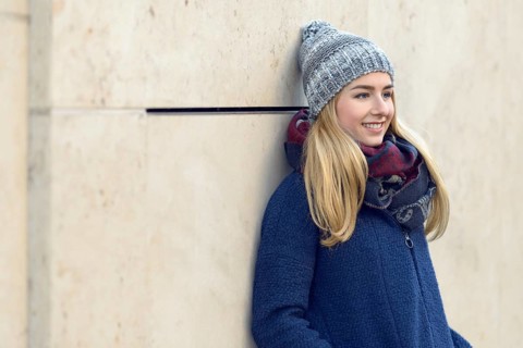 Smiling attractive young blond woman in a warm blue winter outfit standing daydreaming leaning back