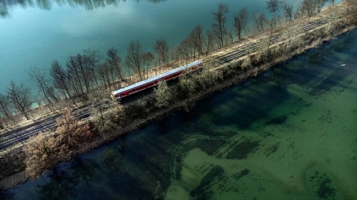 A red local railway runs on an embankment between two turquoise-coloured waters, drone shot, Upper