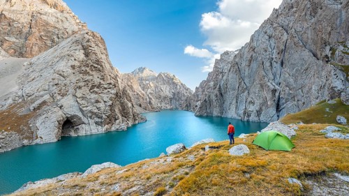 Young man with green tent at the turquoise mountain lake Kol Suu with rocky steep mountains, Kol