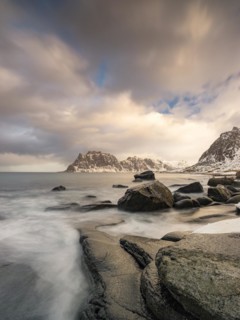 Rounded rocks on Utakleiv beach in a dramatic cloudy atmosphere, snow-capped mountains in the