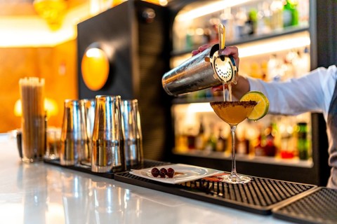 Hand of a waiter filling cocktail into martini glass garnished with lemon in the counter of a