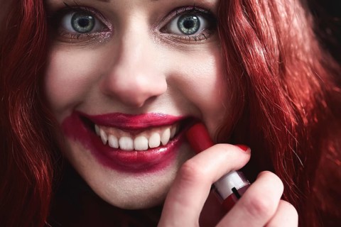 Portrait of crazy-looking teen girl with red hair she is smearing red lipstick on her face, horror