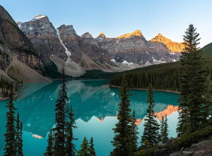 Sunset over a serene lake surrounded by rocky mountains and pine trees, with reflections in the