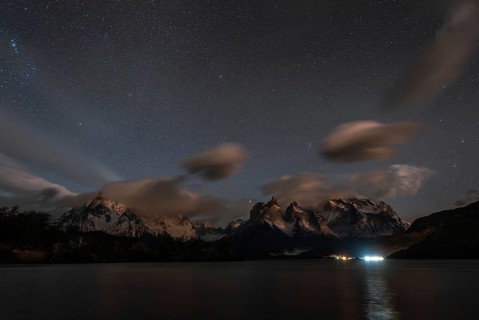 Night sky filled with stars over snow-capped mountains and a calm lake, with clouds and reflections