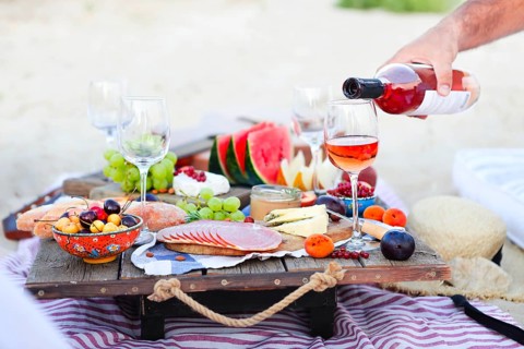 Crop male serving pink wine having picnic with delicious snacks and fruit on sandy beach