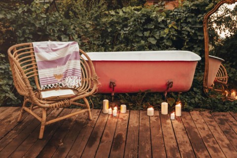 Burning candles and wicker chair with book and wine placed on wooden path near bathtub and mirror