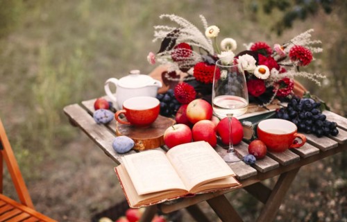 Opened book and glass of wine placed near dishware and bouquet of fresh flowers amidst assorted