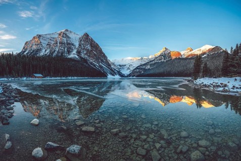 Peaceful lake reflecting snow-capped mountains and a cabin under a clear blue sky at sunrise, Lake
