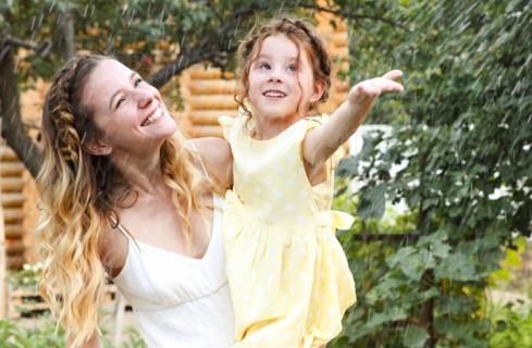 Beautiful happy family of young smiling mother and little daughter under summer rain, having fun