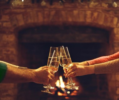People clinking champagne glasses while gathering around fireplace in country house, cropped shot.