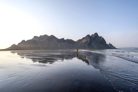 Tourist at black lava beach, sandy beach and sea, mountains Klifatindur reflected in the water,