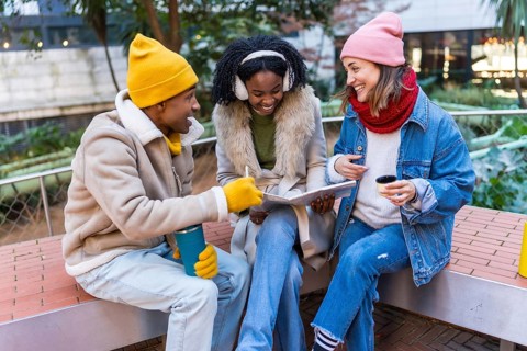 Three cheerful young students are sitting on a bench, studying and having a coffee break outdoors