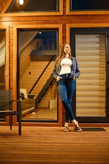 A beautiful brunette woman stands on the doorway of a large wooden cabin in the evening