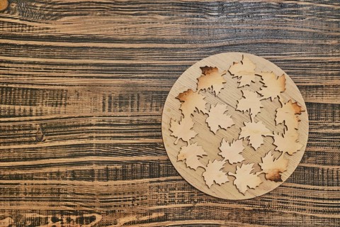 A circular wooden base hot pad displays intricately cut wooden leaves with varying hues, arranged