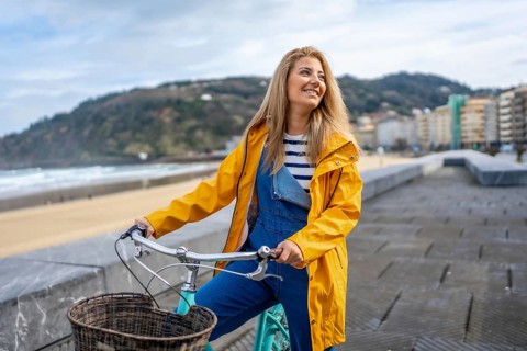 Young woman wearing yellow raincoat riding bicycle on boardwalk by the beach, enjoying ocean view