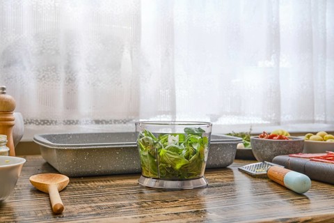 Fresh greens are placed in a chopping bowl of immersion blender on a wooden countertop alongside