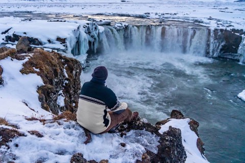 A man hiker sitting looking at the frozen Godafoss waterfall at sunset in winter, Iceland, Europe