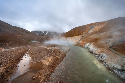 Steaming stream between colourful rhyolite mountains with snowfields, Hveradalir geothermal area,