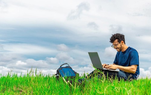 Smiling man with laptop sitting on the grass in the field. Side view of man sitting on green grass