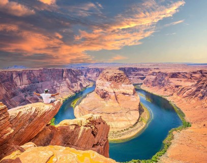 A young man sitting with a white shirt and green pants on Horseshoe Bend and the Colorado River in