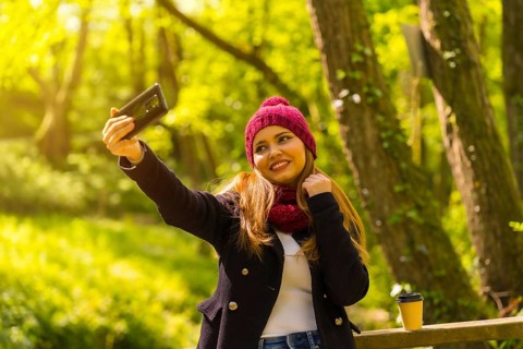 Young man with a black jacket, scarf and red woolen hat enjoying in an autumn park, taking a selfie
