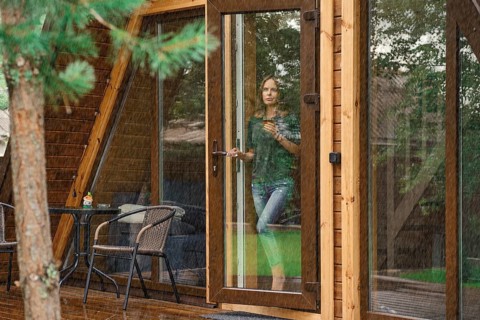 Young woman with a cup of herbal tea stands on the doorstep of a forest cabin and looks at the rain