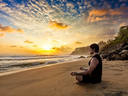 Young sporty fit man doing yoga meditating in padmasana lotus pose on tropical beach on sunset