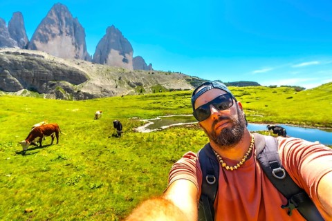 Backpacker taking a selfie with cows grazing in a green meadow in the tre cime di lavaredo national