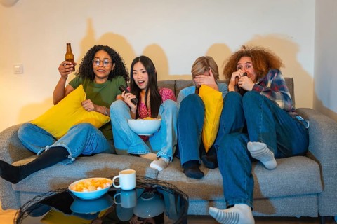 Four friends experiencing different emotions while watching a suspenseful movie at home, enjoying