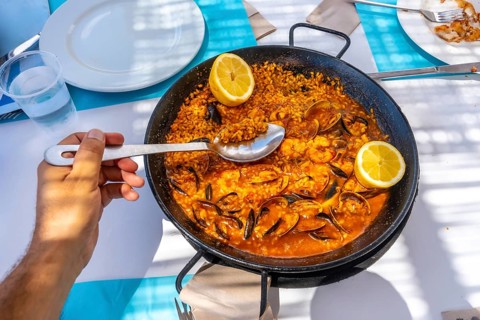 Hand holding spoon scooping seafood paella from pan in outdoor restaurant during summer vacation