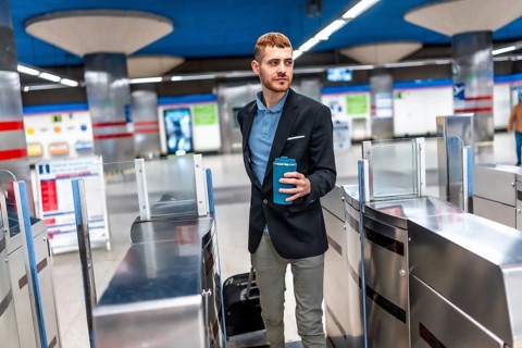 Young businessman going to work passing through subway turnstile holding reusable coffee cup and