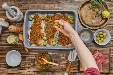 A hand adds green olives to a tray of seasoned fish fillets covered in vibrant herbs and spices.