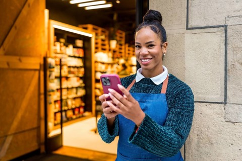 Happy african american saleswoman wearing apron using smartphone at organic supermarket entrance