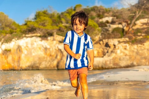 A young boy is running on the beach wearing a striped shirt and shorts. The image has a light and